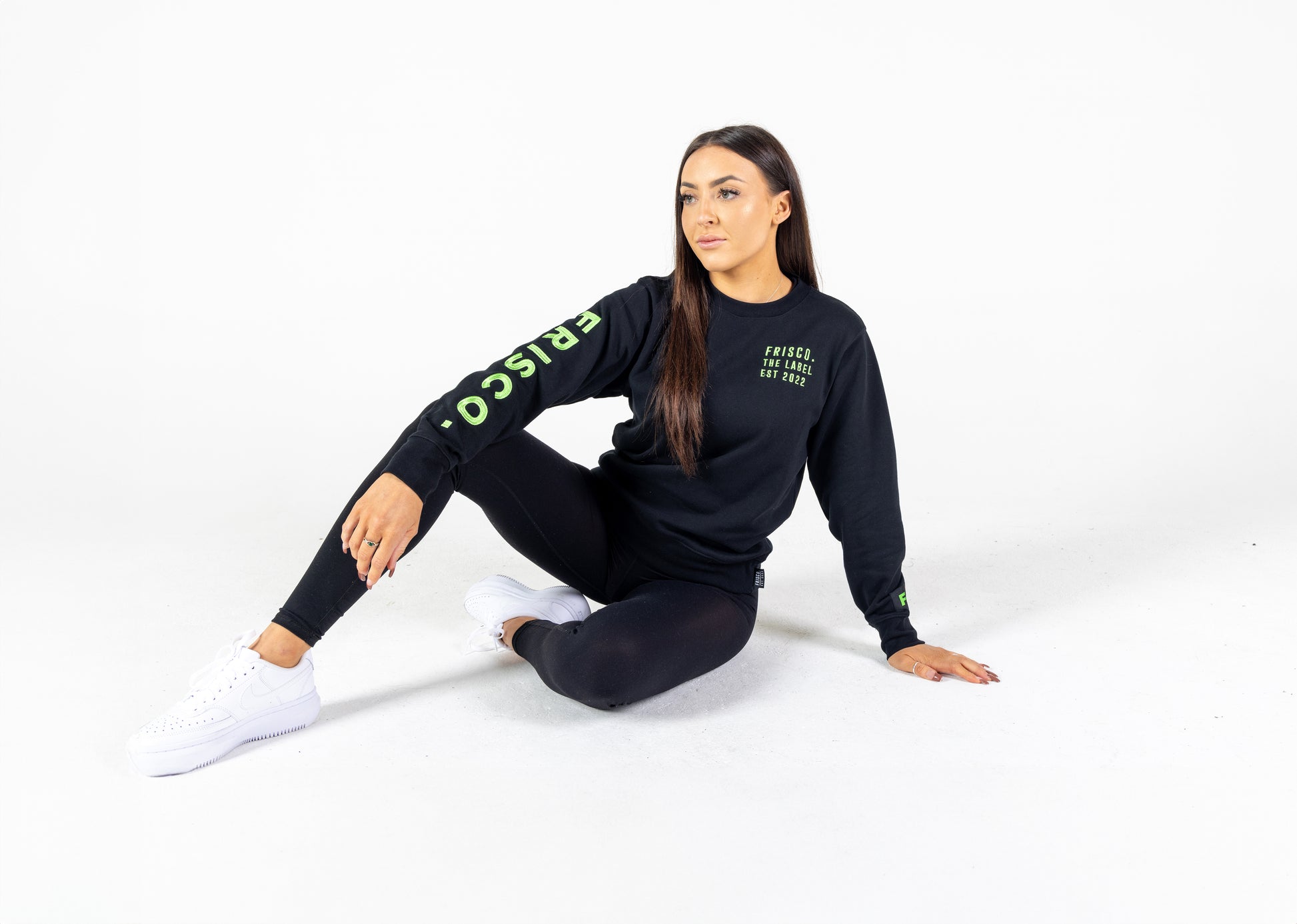 "Young woman seated, modelling Frisco Women's activewear small longsleeve crew top in black with green embroidery "