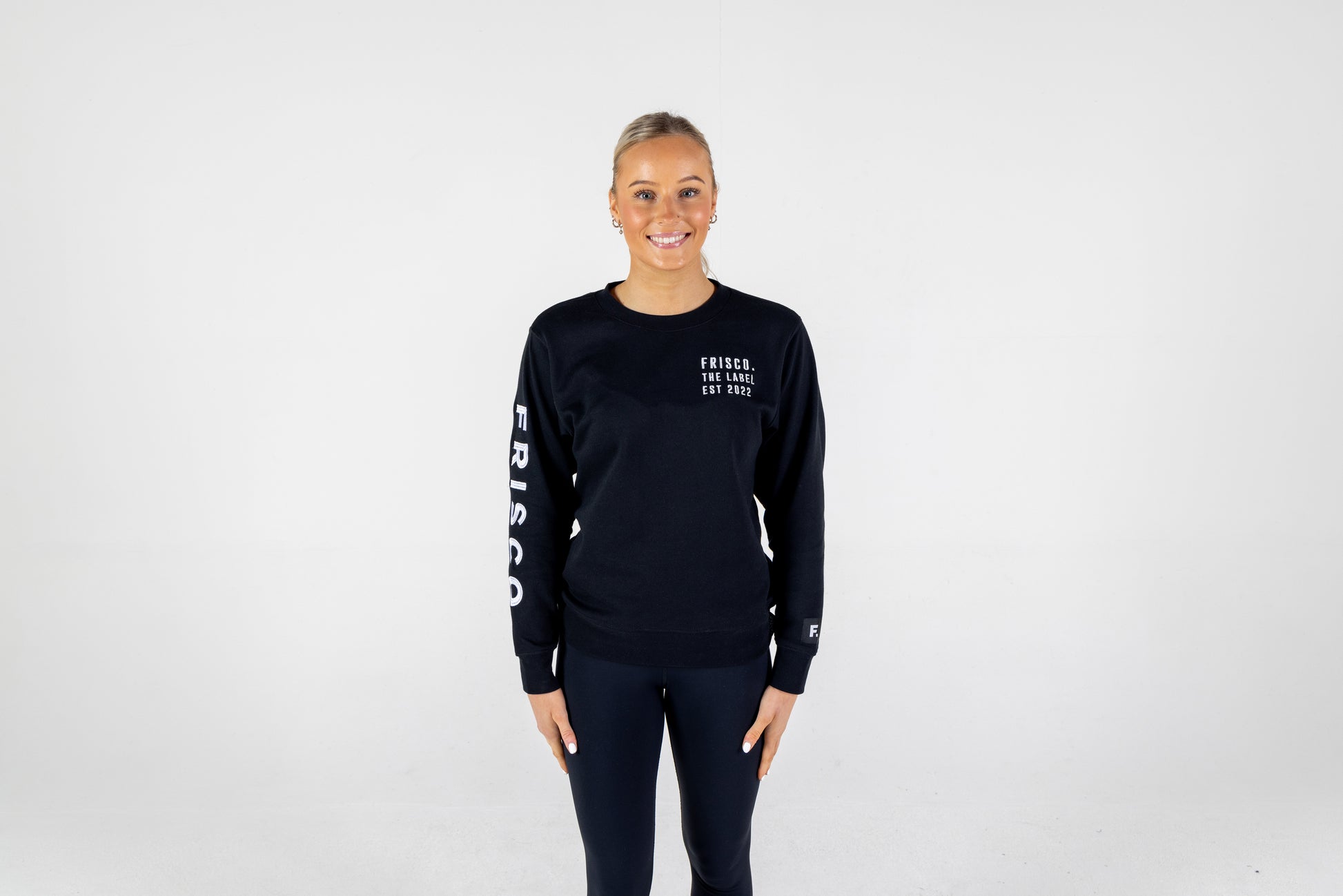 Frisco Women's activewear small longsleeve crew top in black with white embroidery, worn by smiling woman, shown from front