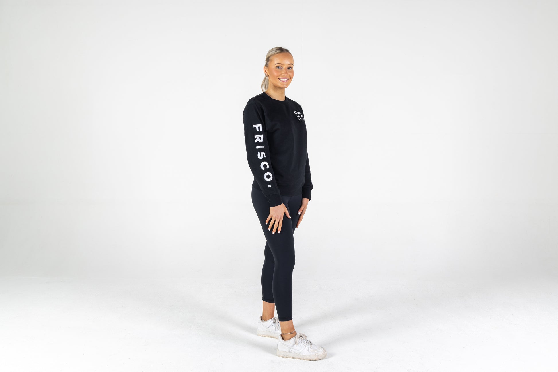 Frisco Women's activewear small longsleeve crew top in black with white embroidery, worn by smiling woman, shown full body from front right side