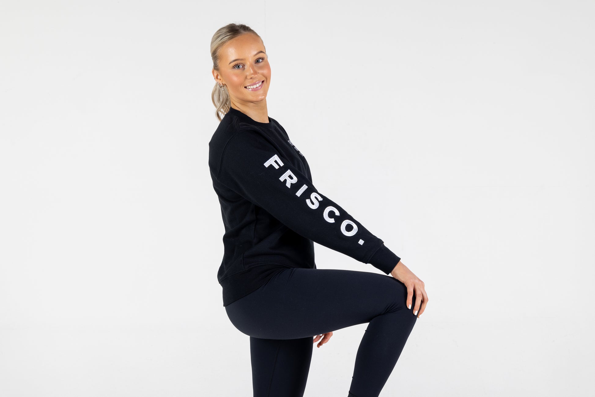 "Frisco Women's activewear small longsleeve crew top in black with white embroidery, worn by smiling woman shown from right side, leg up     "